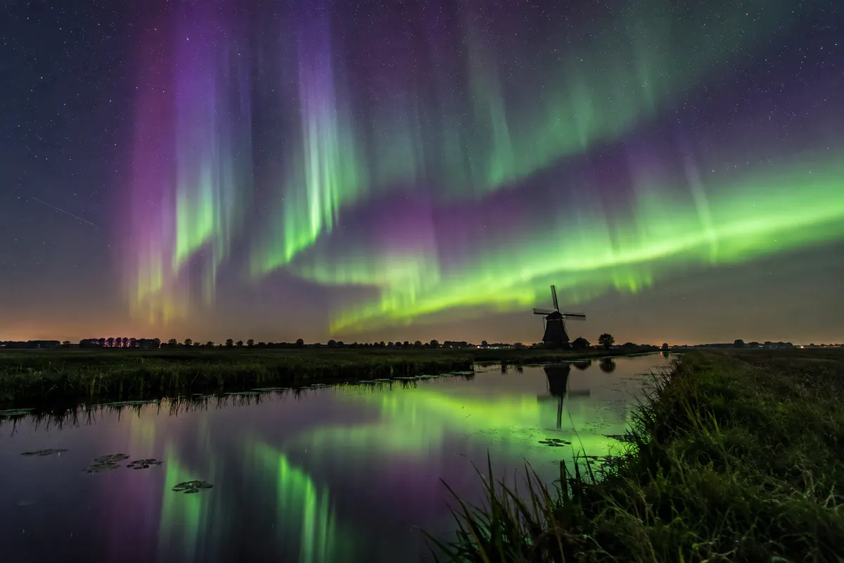 Groene en paarse noorderlichtstralen boven kanaal met windmolen en weerspiegeling